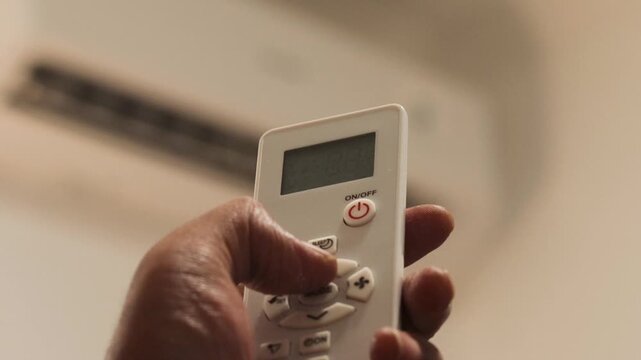 Asian man's hand sets the air conditioner temperature with a remote control in a bedroom before going to sleep during summer. Home cooling and nighttime comfort.
