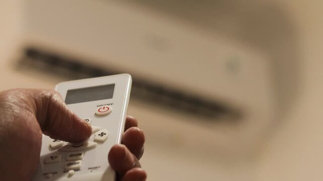 Asian man's hand sets the air conditioner temperature with a remote control in a bedroom before going to sleep during summer. Home cooling and nighttime comfort.