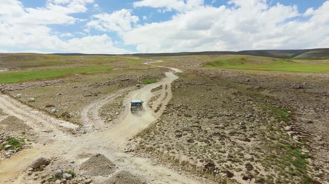 Drone shot following a rugged 4x4 vehicle driving along a bumpy dirt road, kicking up dust trails across the vast, high altitude Deosai Plains under a blue summer sky