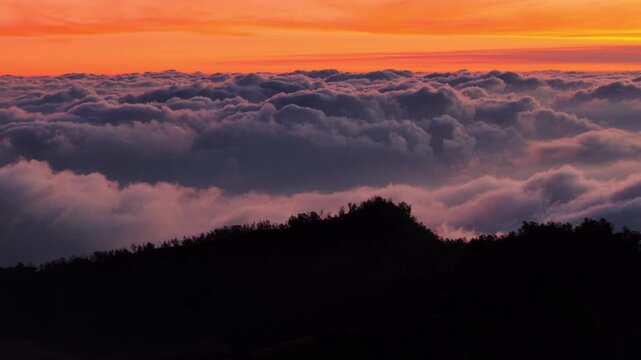 Aerial view of Dieng Plateau with silhouettes of mountain ridges rising above a thick layer of clouds during a vibrant orange sunrise in Indonesia.