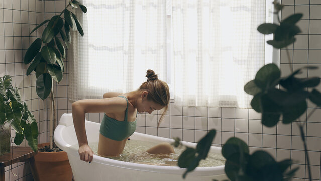 Young Asian woman relaxes in an ice bath in a bright, plant-filled bathroom with white grid tiles, highlighting cold therapy and self-care.
