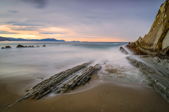Long exposure view of La Salvaje beach in Sopelana featuring rocky flysch formations silhouetted cliffs and a misty sea at sunset