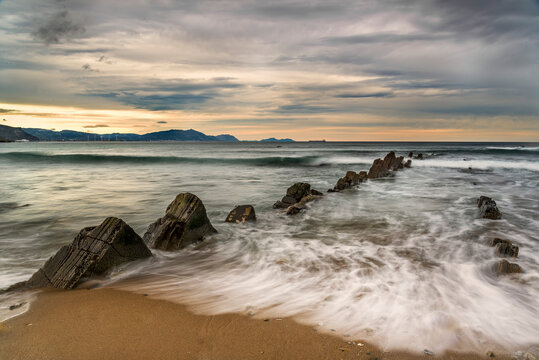 Long exposure view of La Salvaje beach in Sopelana featuring rocky flysch formations silhouetted cliffs and a misty sea at sunset