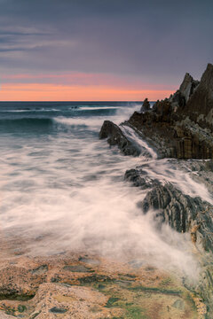 Long exposure view of La Salvaje beach in Sopelana featuring rocky flysch formations silhouetted cliffs and a misty sea at sunset