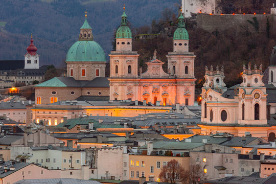 Historic churches and rooftops of Salzburg Old Town illuminated at dusk with wooded hills in the background, Austria.