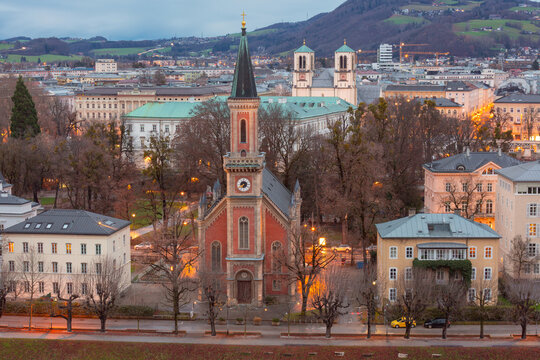 Christ Church surrounded by historic buildings and trees in Salzburg at dusk, Austria, with city districts and hills in the background.