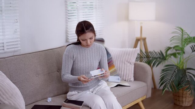 Young Asian woman reviewing furniture design samples on digital tablet while sitting on modern sofa in bright living room