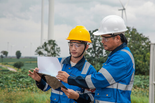 Two engineers in hard hats and safety gear reviewing documents on a clipboard at a wind turbine farm.