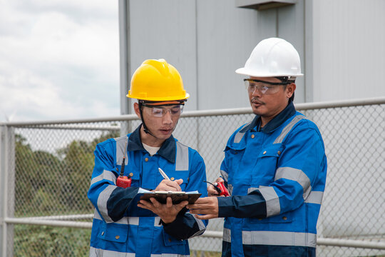 Two engineers in hard hats and safety gear reviewing documents on a clipboard at a wind turbine farm.