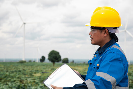 Engineer in a yellow hard hat talking on a walkie-talkie at a wind turbine farm site.