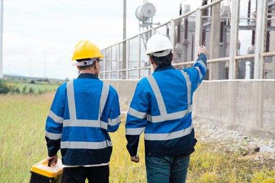 Two engineers in hard hats and safety gear reviewing documents on a clipboard at a wind turbine farm.