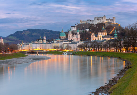 Panoramic view of Salzburg Old Town along the Salzach River with historic churches and Hohensalzburg Fortress illuminated at dusk, Austria.