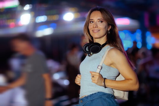 Smiling Young Woman With Headphones Around Neck Posing At Carnival. Attractive cheerful young lady in knit top standing in front of vibrant urban scenery