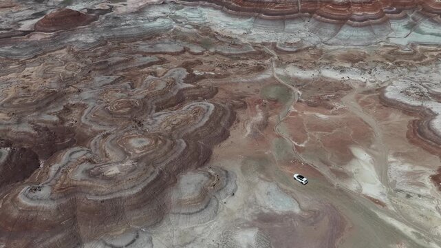 Aerial view of the colorful striped Bentonite Hills with a white car driving along a dirt path in the desert landscape in Hanksville, Utah, United States.