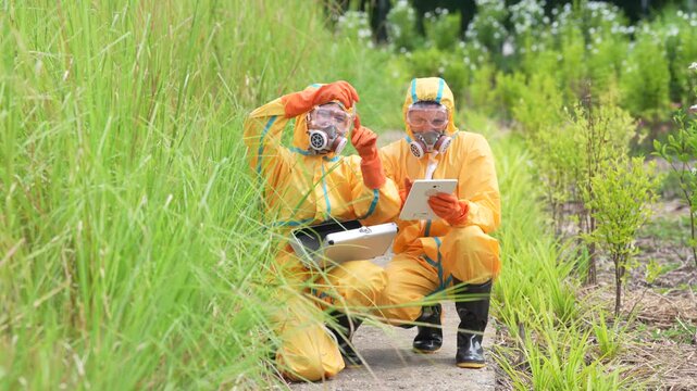 Hazmat suit respirator scientist fieldwork environmental sampling contamination protective glove tablet, researchers kneel on rural roadside grass, collect soil samples, record data on clipboard
