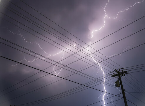 Powerful lightning bolt striking behind power lines, dramatic stormy sky, dangerous electricity, thunderstorm atmosphere, electric pole silhouette