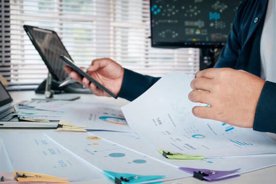 Business professional reviewing financial reports and data analytics on smartphone while examining printed charts and documents at a modern office desk, supporting financial planning, decision making.
