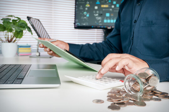 Business professional calculating personal finances with a calculator while holding documents beside a laptop and coins, representing budgeting, expense tracking, savings planning and money management