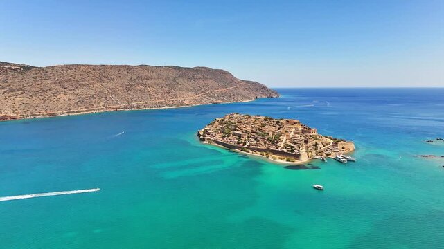 Kalydon fortress island recedes in a wide aerial view off Crete as a speedboat crosses turquoise water, leaving a white wake near docked ferries and an arid headland.