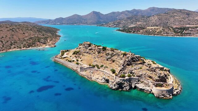 Kalydon stone bastion and perimeter walls extend around a rocky island during an aerial orbit off Crete, surrounded by turquoise channels and arid coastal mountains in clear light.