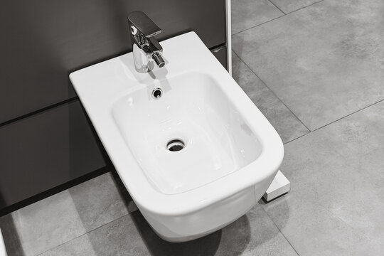 White ceramic bidet with shiny chrome faucet in modern bathroom with a grey tiled floor
