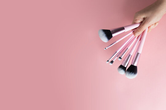 A hand holds set of various pink-handled makeup brushes, fanned out against a soft pink backdrop, ready for flawless application.