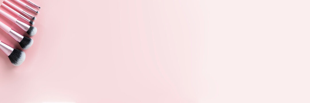 Banner size image showing the handles of several pink makeup brushes lined up on the left side, on a muted pink background with copy space.