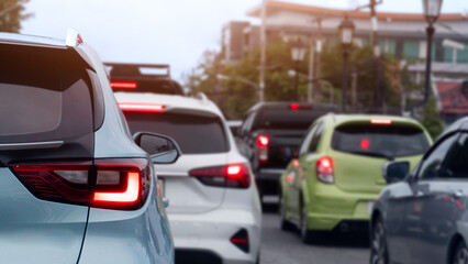 Fototapeta premium Cars wait in a heavy traffic queue on an urban street. During a busy daytime commute with glowing brake lights. Blurred of trees and town under bright sky.