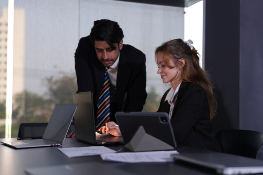 Manager in a colorful striped tie studies the laptop over his colleague's desk while the ginger woman keeps her eyes on her own tablet screen in soft daylight in a bright modern coworking.
