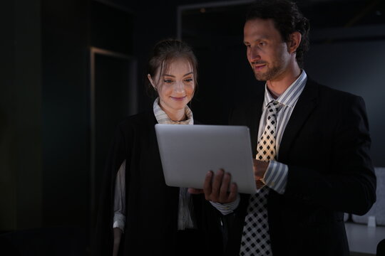 Bearded businessman peers down into the laptop from his full height while the young woman angles the screen up toward him in the pitch dark office in a bright modern coworking office.