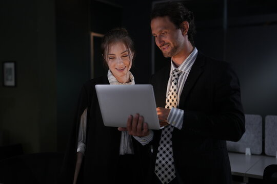 Young woman with a pale scarf laughs at the laptop she carries while the bearded man beside her smiles at the screen in the pitch black office in a bright modern coworking office.