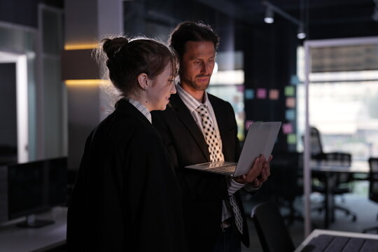 Standing pair reviews the glowing laptop in matched profile, her earring sparking in the screen light as the night office sleeps around them in a bright modern coworking office.