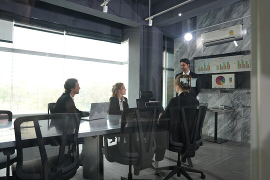Presenter turns to point out a figure on the wall screen while his three colleagues study the charts from their seats in the sun-washed marble conference room in a bright modern coworking.