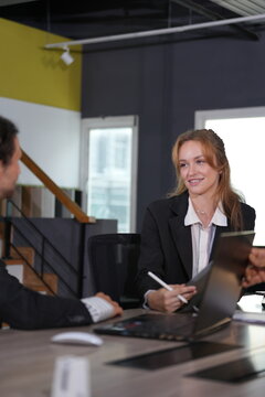 Grinning red-haired professional holds her stylus up while chatting over the open laptop, her colleague's arm resting on the desk in the yellow-walled loft in a bright modern coworking.