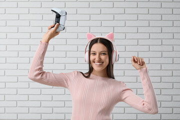 Gaming culture and entertainment, happy young woman with cat ear headphones holding a game controller in front of a white brick wall © Pixel-Shot