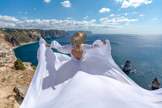 Woman dress cliff, elegant lady in flowing white gown standing on rocky coast overlooking beautiful blue sea with copy space