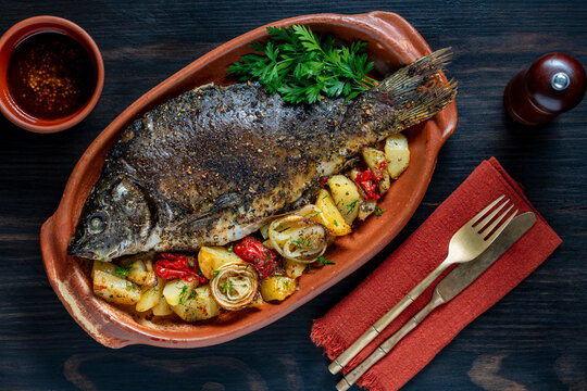 Baked carp fish with potatoes, tomatoes, and onions. Flat lay food photography for seafood restaurant menu, healthy recipe, or dinner concept.