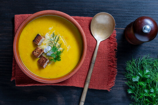 Pumpkin cream soup with croutons and herbs closeup. Creamy yellow vegetable soup with croutons, cheese, and fresh herbs in bowl. Rustic flat lay food concept for autumn menu, recipe, and restaurant