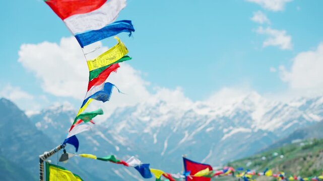 4K Slow motion shot of snowy Himalayan mountain range with blue sky and clouds with defocused colorful Buddhist Tibetan prayer flags waving in the wind in front of it at Lahaul in Himachal Pradesh.