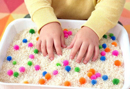 Child playing with colorful pom poms in a sensory bin filled with rice for creative learning and fun