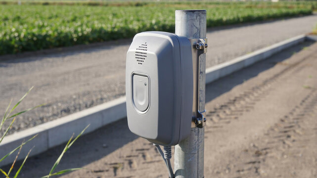 Gray outdoor emergency call box or speaker device mounted on a metal pole next to a road and green field