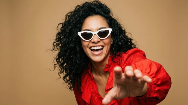 Cheerful Young Woman with Curly Hair Wearing White Sunglasses and Red Blouse Reaching Towards Camera on Beige Background