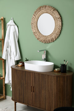 Modern sink with bath supplies on countertop and stylish mirror near green wall in bathroom