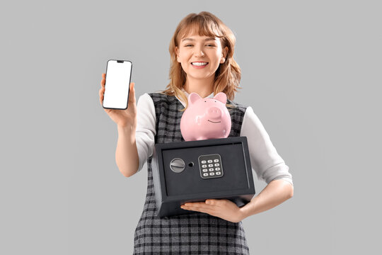 Beautiful young happy woman with safe box, piggy bank and mobile phone on grey background