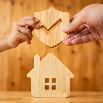 Family home insurance and property protection concept with adult and child hands holding wooden shield with checkmark over house model for mortgage safety and residential coverage.