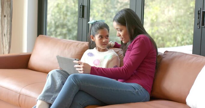 Daughter asking question triggers mother leaning and tapping tablet guiding on sofa in living room