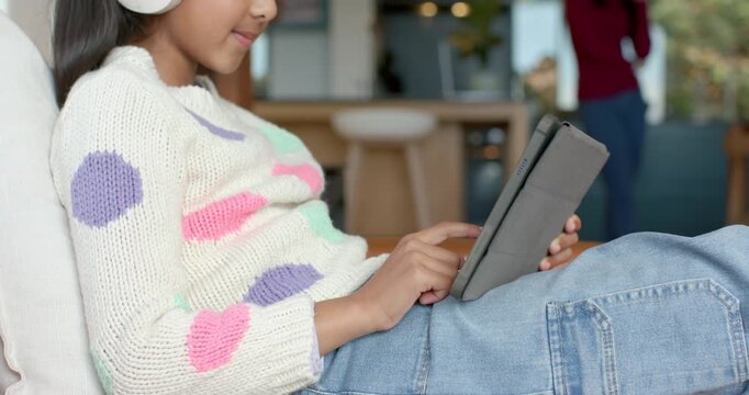 Indian girl putting on white headphones and tapping tablet while mother talking on phone walking by