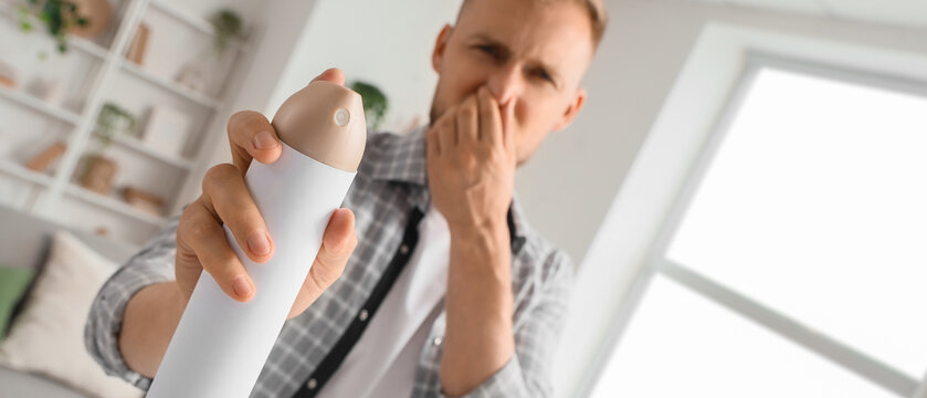 Bad odor and air freshener usage, man covering nose while spraying aerosol to remove unpleasant smell in home living room environment