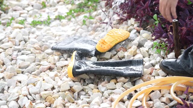 Garden irrigation hose water spray on gravel, rubber boot outdoor footwear rinsing with stream and sponge, splashing yard care cleanup on stone path, practical cleaning during daylight work