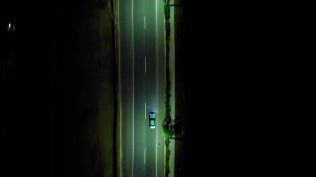 Top-down zenith aerial drone shot tracking a patrol vehicle with flashing emergency lights driving along an illuminated multi-lane highway at night